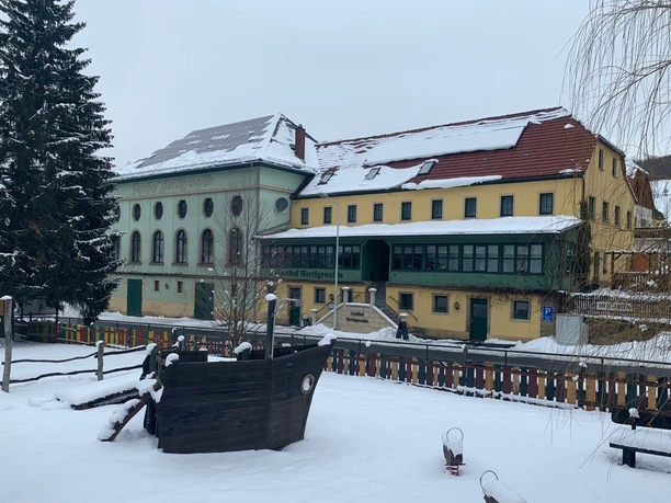 A snow-covered playground with a wooden ship in the foreground, behind it a large, yellow-green building with red roofs and a Christmas tree.