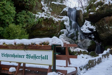 Besneeuwde Lichtenhain waterval met bevroren watervallen, omgeven door groene planten en rotsen; een bord geeft de locatie aan.