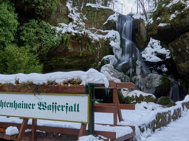 Snow-covered Lichtenhain waterfall with frozen cascades, surrounded by green plants and rocks; a sign indicates the location.