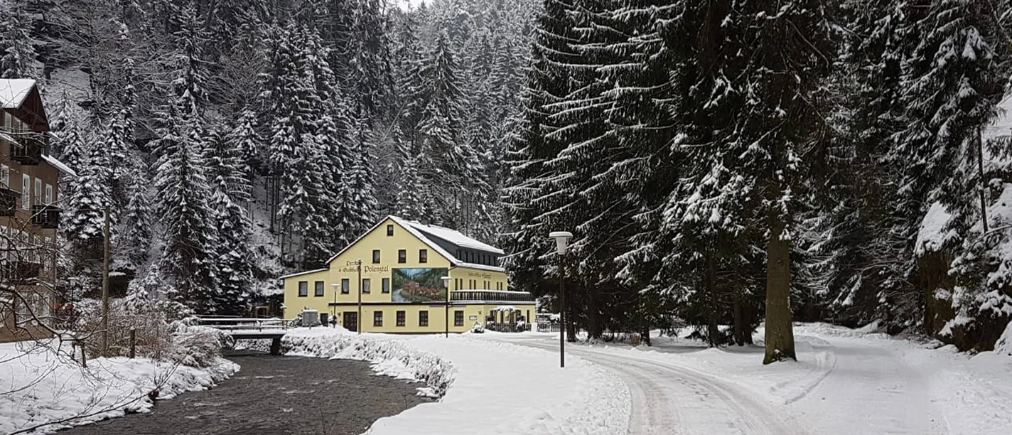 Žlutá budova v zasněžené lesní krajině, vedle potoka a zasněžené cesty, obklopená vysokými jedlemi.