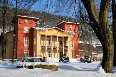 Parkhotel Bad Schandau Winter A large, two-tone building in red and yellow stands in a snowy landscape with trees and a blue sky in the background.
