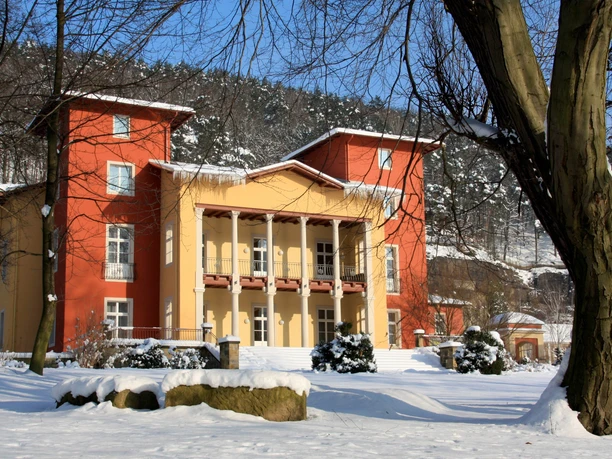 Parkhotel Bad Schandau Winter V zasněžené krajině se stromy a modrou oblohou v pozadí stojí velká dvoubarevná budova v červené a žluté barvě.