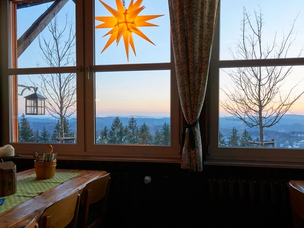 Bekijk Wachbergbaude Houten tafel met kaars en bestek voor een raam met uitzicht op besneeuwde bomen en bergen bij zonsondergang; een stralende ster hangt in het raam.