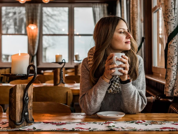 Gastraum Wachbergbaude Eine Frau in einem gemütlichen Café hält eine Tasse, schaut aus dem Fenster; auf dem Tisch brennt eine Kerze, warme Atmosphäre.