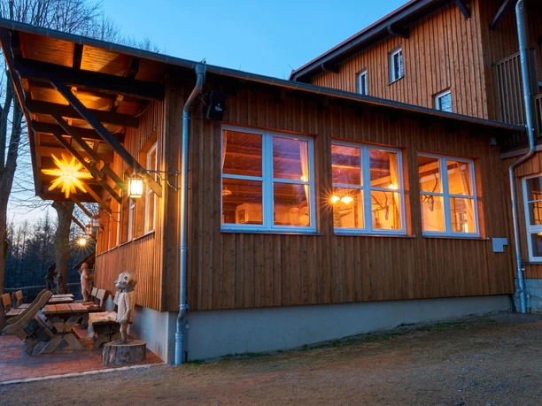 Wachbergbaude Holzhaus mit großen Fenstern, warmem Licht und leuchtendem Stern an der Fassade; rustikale Sitzbänke und Holzfigur im Vordergrund, abendliche Stimmung.
