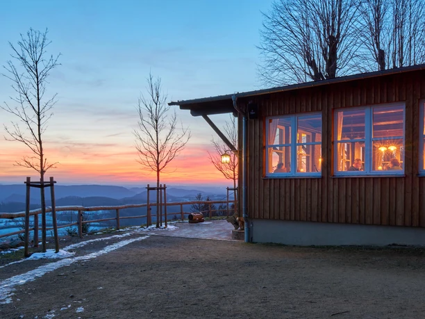 Wachbergbaude Holzhütte mit erleuchteten Fenstern bei Sonnenuntergang, umgeben von kahlen Bäumen und einem verschneiten Weg, mit Blick auf hügelige Landschaft.