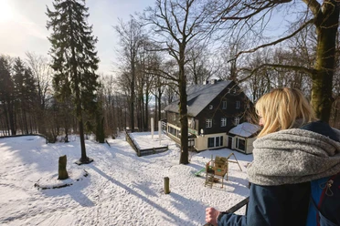 Ungerberg Ausflugsgaststätte & Pension Eine Frau mit blonden Haaren und grauem Schal blickt auf ein verschneites Haus im Wald; die Sonne scheint, die Atmosphäre ist ruhig.