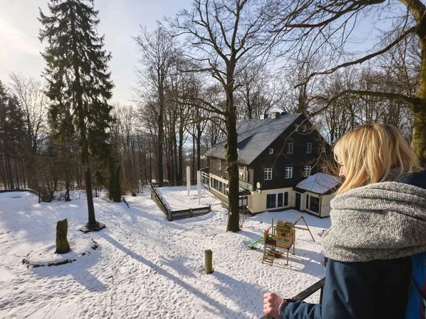Ungerberg excursion restaurant & guesthouse A woman with blonde hair and a gray scarf looks at a snow-covered house in the forest; the sun is shining, the atmosphere is calm.