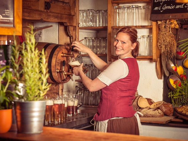 "Restauratie vesting Königstein "Zum Musketier Een vrouw in een rood vest tapt bier uit een houten vat in een rustieke bar; glazen en verse groenten zijn te zien op de achtergrond.
