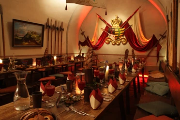 Casemates Gastronomy Experience Restaurant Königstein Fortress Rustic dining room with long wooden tables, decorated with candles, red napkins and bread baskets; a coat of arms and halberds on the wall.