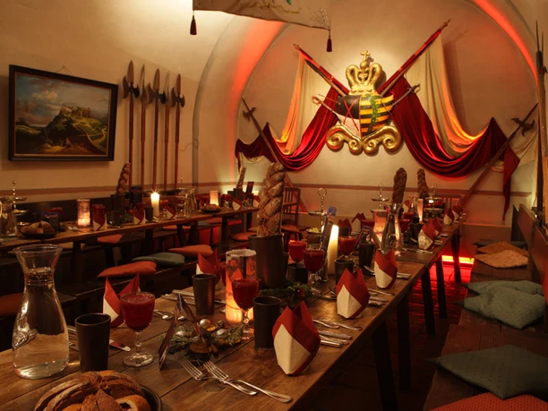 Zážitková gastronomická restaurace Casemates Pevnost Königstein Rustikální jídelna s dlouhými dřevěnými stoly, zdobená svíčkami, červenými ubrousky a košíky na chléb; na stěně erb a halapartny.