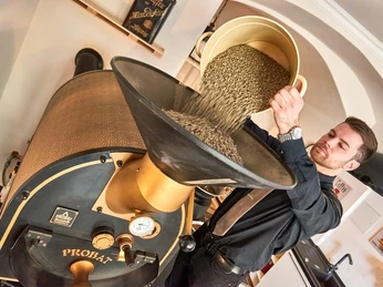 Café & Weinhaus Kaffeerösterei Schmole in Pirna Ein Mann in schwarzer Kleidung schüttet grüne Kaffeebohnen aus einem gelben Behälter in eine goldene Kaffeeröstmaschine.