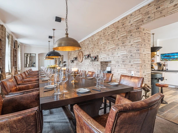 Café & Wine House Pražírna kávy Schmole v Pirně Elegantní jídelna s dlouhým dřevěným stolem, prostřeným sklenicemi a svíčkami, obklopená hnědými koženými křesly a osvětlená velkými závěsnými lampami.