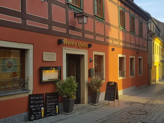 Fachwerkhaus mit roter Fassade und grünen Fensterläden, beschriftet als "Rotes Haus", mit Menütafeln und Pflanzen vor der Tür.Half-timbered house with red façade and green shutters, labeled "Red House", with menu boards and plants in front of the door.Hrázděný dům s červenou fasádou a zelenými okenicemi, označený jako "Red House", s jídelními lístky a rostlinami přede dveřmi.Dom z muru pruskiego z czerwoną fasadą i zielonymi okiennicami, oznaczony jako "Red House", z tablicami z menu i roślinami przed drzwiami.Vakwerkhuis met rode gevel en groene luiken, met het opschrift "Red House", met menuborden en planten voor de deur.Casa a graticcio con facciata rossa e persiane verdi, etichettata come "Casa Rossa", con cartelli per il menu e piante davanti alla porta.