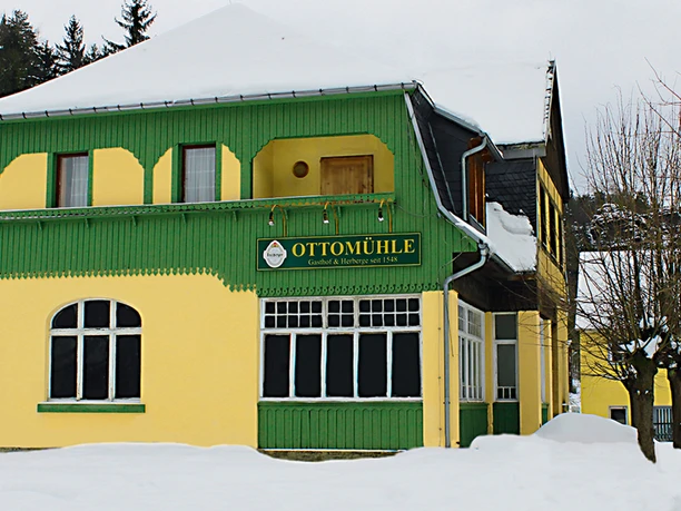 Otto mill in winter Yellow building with green wooden decor and the inscription "OTTOMÜHLE" in a snowy winter, surrounded by bare trees.