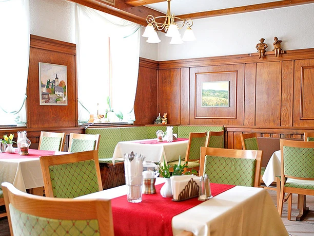Gezellig restaurant met houten muren, groene stoelen en rode tafellopers; lichte sfeer dankzij grote ramen en witte gordijnen.