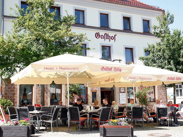 Buitenruimte van een herberg met beige parasols en verschillende mensen aan tafels; ontspannen sfeer bij zonnig weer.