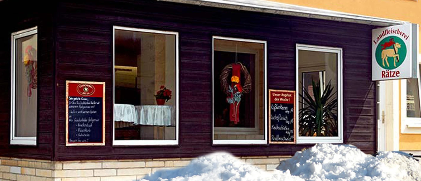 Butcher's shop with dark brown façade, shop windows, signs and piles of snow in front of it. One sign shows the logo of Landfleischerei Rätze.