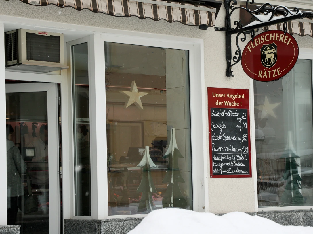 Eingang einer Fleischerei mit rotem Schild und Wochenangebot auf einer Tafel; Fenster mit Stern- und Tannenbaumdekoration, Schnee im Vordergrund.Entrance to a butcher's shop with red sign and weekly specials on a blackboard; window with star and Christmas tree decorations, snow in the foreground.Vchod do řeznictví s červeným nápisem a týdenními nabídkami na tabuli; výloha s hvězdou a ozdobami vánočního stromku, v popředí sníh.Wejście do sklepu mięsnego z czerwonym szyldem i cotygodniowymi promocjami na tablicy; okno z gwiazdą i dekoracjami choinkowymi, śnieg na pierwszym planie.Ingang van een slagerij met rood bord en wekelijkse specials op een schoolbord; raam met ster en kerstboomversiering, sneeuw op de voorgrond.Ingresso di una macelleria con insegna rossa e specialità settimanali su una lavagna; vetrina con stelle e decorazioni per l'albero di Natale, neve in primo piano.