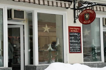 Fleischerei Rätze Eingang einer Fleischerei mit rotem Schild und Wochenangebot auf einer Tafel; Fenster mit Stern- und Tannenbaumdekoration, Schnee im Vordergrund.