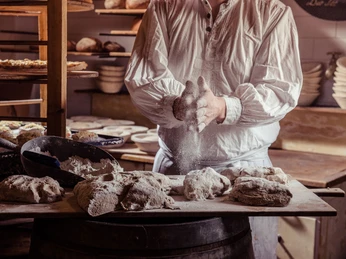 Bio-Bäckerei in Schmilka Bäcker in weißem Hemd knetet Teig auf einem Holztisch, umgeben von Mehl und Brotlaiben; rustikale Backstube mit warmem Licht.Baker in white shirt kneading dough on a wooden table, surrounded by flour and loaves of bread; rustic bakery with warm light.Pekař v bílé košili hněte těsto na dřevěném stole, obklopen moukou a bochníky chleba; rustikální pekárna s teplým světlem.Piekarz w białej koszuli ugniatający ciasto na drewnianym stole, otoczony mąką i bochenkami chleba; rustykalna piekarnia z ciepłym światłem.Bakker in wit overhemd kneedt deeg op een houten tafel, omringd door bloem en broden; rustieke bakkerij met warm licht.Fornaio in camicia bianca che impasta su un tavolo di legno, circondato da farina e pagnotte; panificio rustico con luce calda.