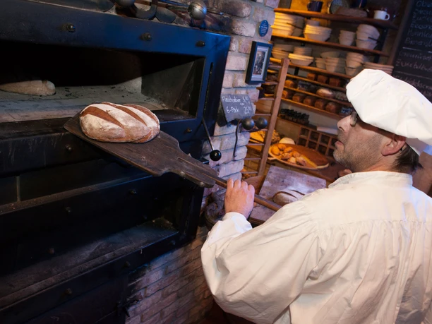 Holzofen in der Bio-Bäckerei in Schmilka Bäcker in weißer Kleidung zieht frisches Brot mit einer Schaufel aus einem traditionellen Backofen; im Hintergrund Regale mit weiteren Broten.