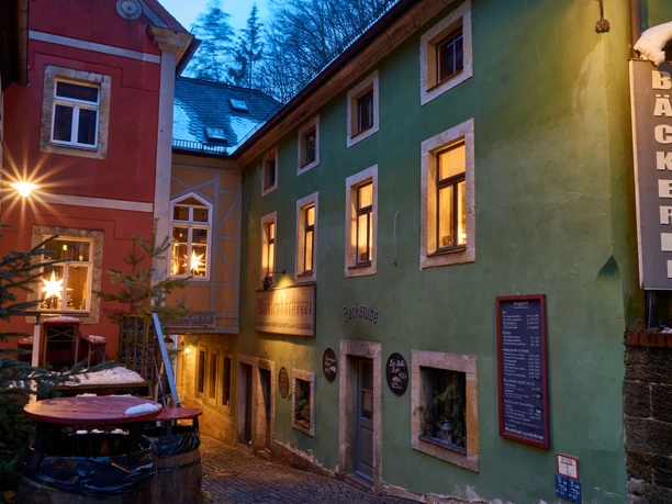Bio-Bäckerei im Winter Enge Gasse mit einer rot-grünen Bäckerei, beleuchtet von warmem Licht, weihnachtliche Dekorationen in den Fenstern, winterliche Atmosphäre.