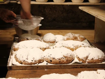 Traditioneller Stollen in der Bio-Bäckerei in Schmilka Bäcker bestäubt frisch gebackene Stollen mit Puderzucker; die Stollen liegen auf einem Backblech in einer rustikalen Bäckerei.Baker dusting freshly baked stollen with powdered sugar; the stollen lie on a baking tray in a rustic bakery.Pekař sype čerstvě upečený štolu moučkovým cukrem; štoly leží na plechu v rustikální pekárně.Piekarz posypujący świeżo upieczone stollen cukrem pudrem; stollen leżą na blasze w rustykalnej piekarni.Bakker bestuift versgebakken stollen met poedersuiker; de stollen liggen op een bakplaat in een rustieke bakkerij.Panettiere che spolvera di zucchero a velo gli stollen appena sfornati; gli stollen giacciono su una teglia in una panetteria rustica.