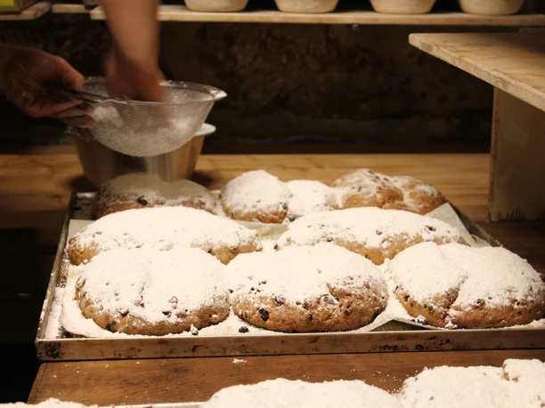 Traditioneller Stollen in der Bio-Bäckerei in Schmilka Piekarz posypujący świeżo upieczone stollen cukrem pudrem; stollen leżą na blasze w rustykalnej piekarni.