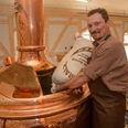 Ein Mann in kariertem Hemd und Schürze schüttet einen Sack Malz in einen großen kupfernen Braukessel in einer Brauerei.A man in a checked shirt and apron pours a bag of malt into a large copper brewing kettle in a brewery.Muž v kostkované košili a zástěře sype pytel sladu do velkého měděného varného kotle v pivovaru.Mężczyzna w kraciastej koszuli i fartuchu wlewa worek słodu do dużego miedzianego kotła w browarze.Een man in een geruit hemd en schort giet een zak mout in een grote koperen brouwketel in een brouwerij.Un uomo in camicia a scacchi e grembiule versa un sacco di malto in un grande bollitore di rame in una fabbrica di birra.