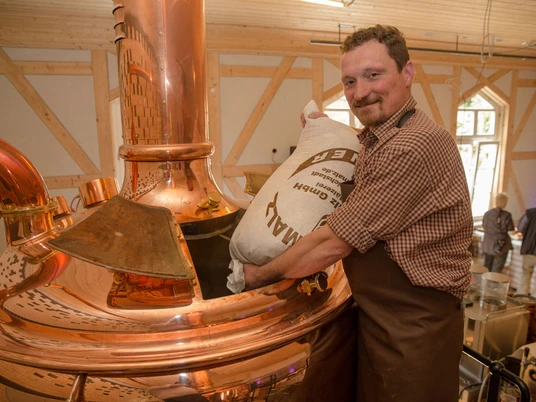 Ein Mann in kariertem Hemd und Schürze schüttet einen Sack Malz in einen großen kupfernen Braukessel in einer Brauerei.A man in a checked shirt and apron pours a bag of malt into a large copper brewing kettle in a brewery.Muž v kostkované košili a zástěře sype pytel sladu do velkého měděného varného kotle v pivovaru.Mężczyzna w kraciastej koszuli i fartuchu wlewa worek słodu do dużego miedzianego kotła w browarze.Een man in een geruit hemd en schort giet een zak mout in een grote koperen brouwketel in een brouwerij.Un uomo in camicia a scacchi e grembiule versa un sacco di malto in un grande bollitore di rame in una fabbrica di birra.