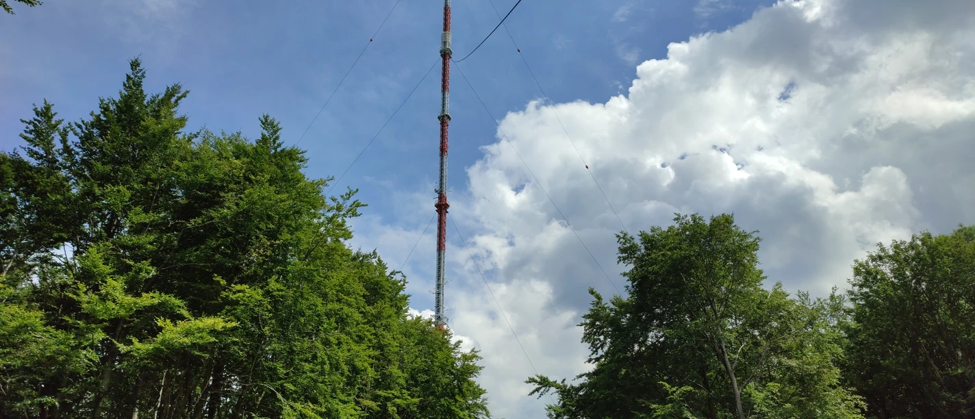 Ein hoher Sendemast ragt über grüne Baumwipfel, umgeben von blauem Himmel und weißen Wolken.