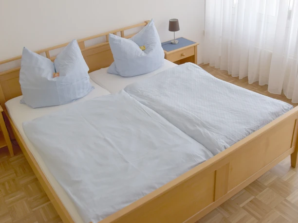 Hotel Im Homburger Ländchen.jpg Doppelbett in einem hellen Raum mit Holzfußboden, blauen Nachttischen und weißen Vorhängen.