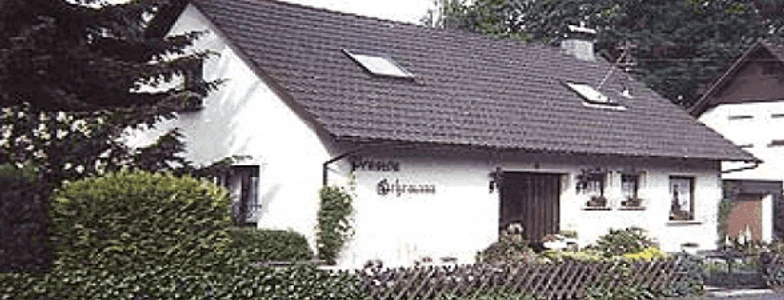 Pension_Gehrmann.jpg.png Ein weißes, einstöckiges Landhaus mit Satteldach, umgeben von grünem Buschwerk und Holzzaun.