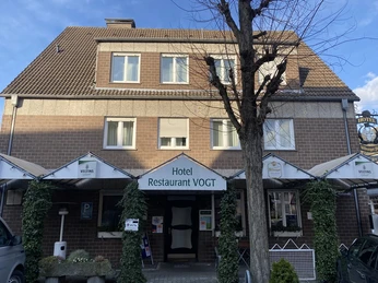 Fassade eines Backsteingebäudes mit dem Schild "Hotel Restaurant VOGT", umgeben von grüner Bewachsung.