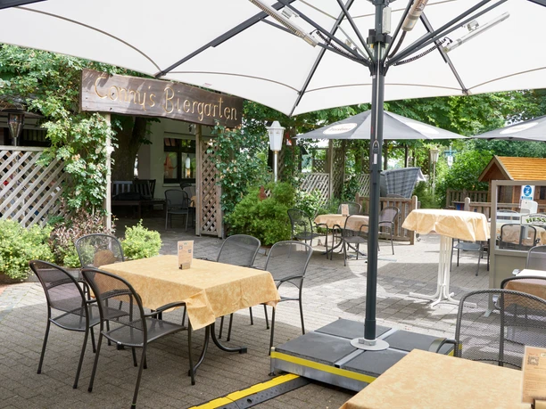 Ein einladender Biergarten mit Tischen, Stühlen, Sonnenschirmen und grünem Laub im Hintergrund.