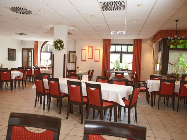 Ein elegantes Restaurant mit roten Stühlen, weißen Tischdecken und großen Fenstern mit Blick ins Grüne.