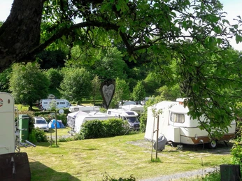 Wohnwagen und Zelte in gepflegter, grüner Umgebung mit Bäumen, Sonnenschein und Herz-Dekoration.