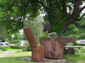 Holztisch und -stühle unter einem Baum auf einem Campingplatz, Wohnwagen im Hintergrund.