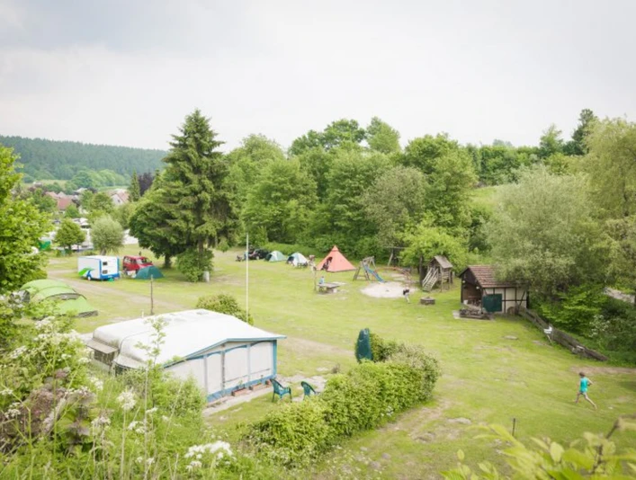Grüner Campingplatz mit Zelten und Wohnwagen, umgeben von Bäumen, mit Spielplatz im Hintergrund.