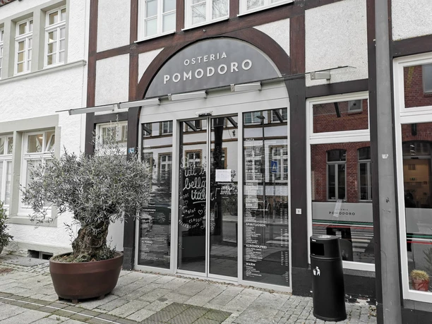 Eingang einer italienischen Osteria mit großen Glasfenstern und Olivenbaum im Topf davor.