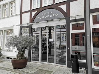 Osteria Pomodoro_Außenansicht Eingang einer italienischen Osteria mit großen Glasfenstern und Olivenbaum im Topf davor.