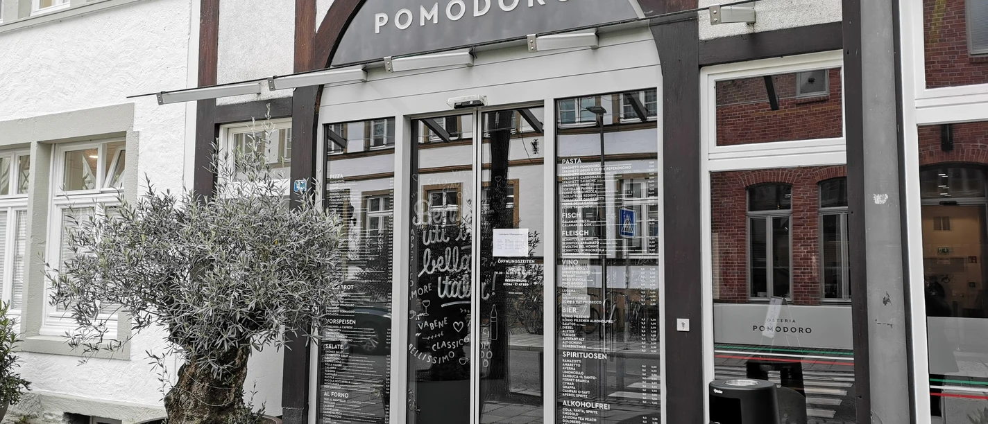 Osteria Pomodoro_Außenansicht Eingang einer italienischen Osteria mit großen Glasfenstern und Olivenbaum im Topf davor.
