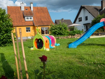 Ferienhaus Westerkamp Garten Spielplatz mit bunter Rutsche und Tunnel auf einer grünen Wiese vor einem Haus mit orangem Dach.