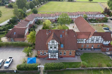Außenansicht Großes Fachwerkgebäude mit roten Dachziegeln und weißen Fensterrahmen, umgeben von begrünten Innenhöfen und parkenden Autos. Der Rehlinger Hof liegt in ruhiger, ländlicher Umgebung mit barrierefrei zugänglichen Außenbereichen.