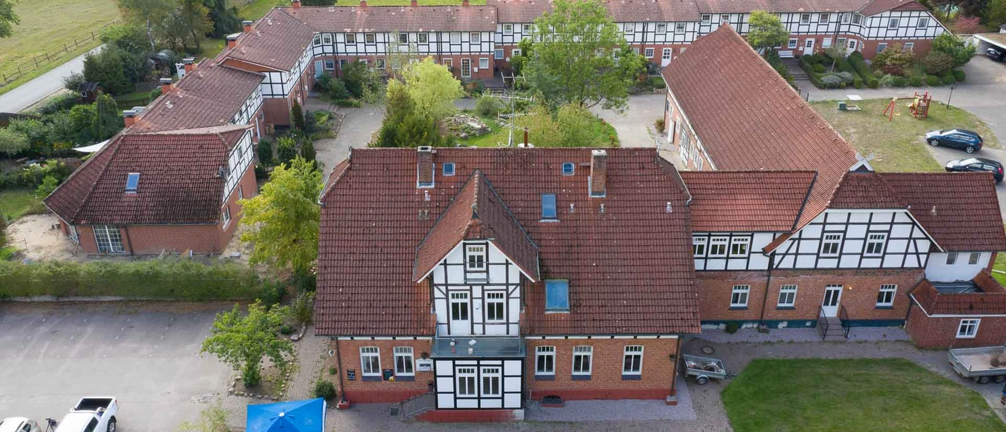 Außenansicht Großes Fachwerkgebäude mit roten Dachziegeln und weißen Fensterrahmen, umgeben von begrünten Innenhöfen und parkenden Autos. Der Rehlinger Hof liegt in ruhiger, ländlicher Umgebung mit barrierefrei zugänglichen Außenbereichen.