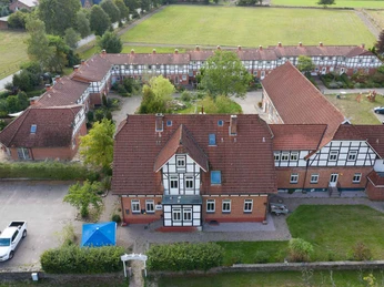 Außenansicht Großes Fachwerkgebäude mit roten Dachziegeln und weißen Fensterrahmen, umgeben von begrünten Innenhöfen und parkenden Autos. Der Rehlinger Hof liegt in ruhiger, ländlicher Umgebung mit barrierefrei zugänglichen Außenbereichen.