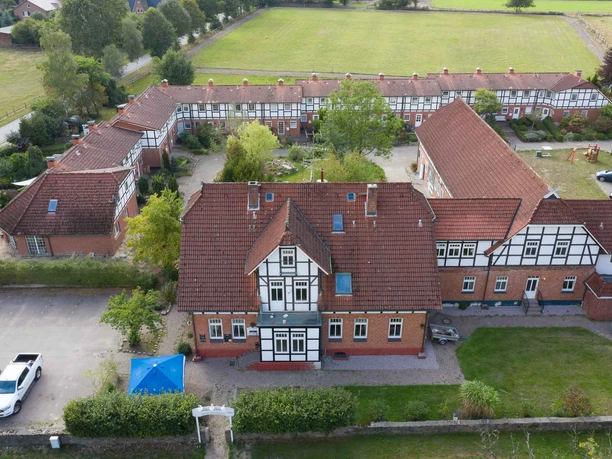 Außenansicht Großes Fachwerkgebäude mit roten Dachziegeln und weißen Fensterrahmen, umgeben von begrünten Innenhöfen und parkenden Autos. Der Rehlinger Hof liegt in ruhiger, ländlicher Umgebung mit barrierefrei zugänglichen Außenbereichen.