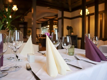 Gastraum Eindeutig gedeckter Tisch mit weißen Tischdecken, gefalteten Stoffservietten in Creme und Lila, mehreren Weingläsern und Besteck. Im Hintergrund Fachwerkstruktur, warme Beleuchtung und grüne Pflanzen. Der Raum wirkt gepflegt und für ein Restaurantessen vorbereitet.