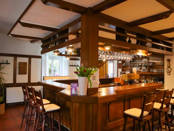 Thekenbereich Holzverkleideter Thekenbereich mit Barhockern und Gläsern über der Theke. Auf der polierten Holzfläche steht eine Vase mit Tulpen. Im Hintergrund sind Regale mit Flaschen und Gläsern, eine helle Wand mit Fenster und eine grüne Pflanze sichtbar.
